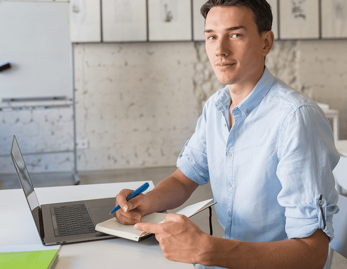 Ein Mann in einem hellblauen Hemd sitzt an einem Schreibtisch mit einem Laptop, hält ein Notizbuch und einen Stift in der Hand und schaut in die Kamera in einer hellen, modernen Büroumgebung.