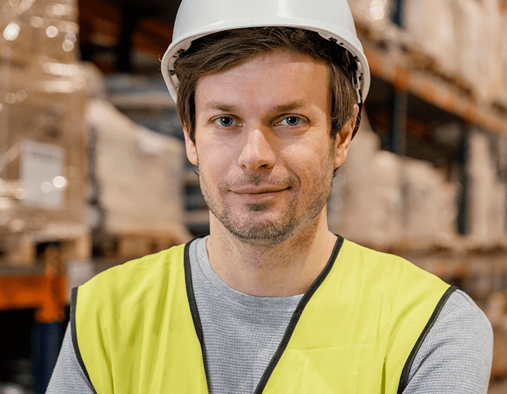Ein Mann mit einem weißen Schutzhelm und einer gelben Sicherheitsweste steht in einem Lagerhaus und schaut in die Kamera. Im unscharfen Hintergrund sind Stapel von Kisten und Regalen zu sehen.