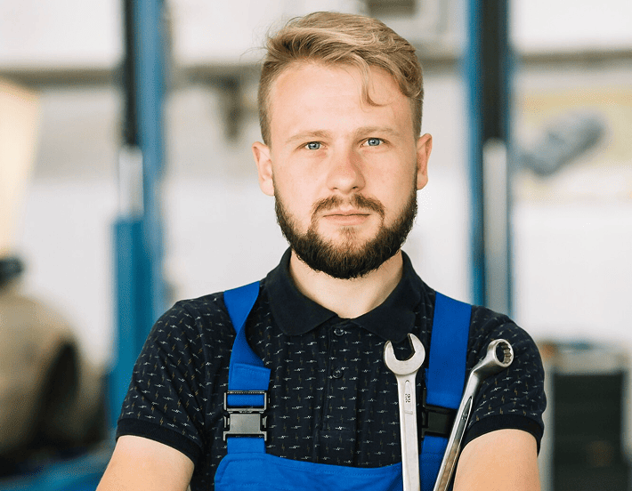 Ein bärtiger Mann in einem blauen Overall und schwarzem Hemd steht in einem Haus und hält zwei Schraubenschlüssel in der Hand. Der Hintergrund ist unscharf.