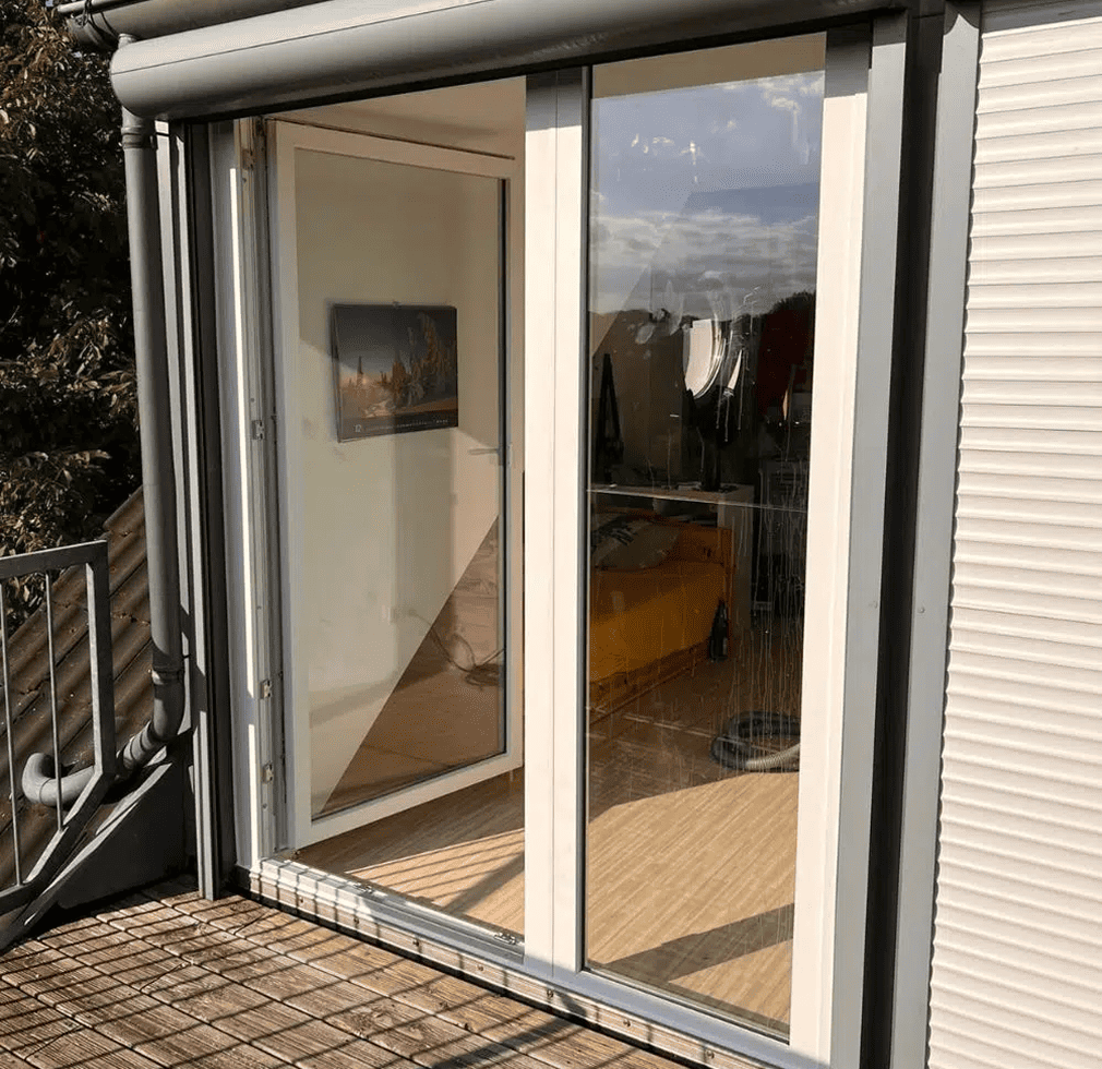 Eine Glastür führt auf einen Balkon mit einem Metallgeländer. In der Tür spiegeln sich der blaue Himmel und die Wolken, und drinnen hängt ein Gemälde an der Wand über einem hellen Holzfußboden.