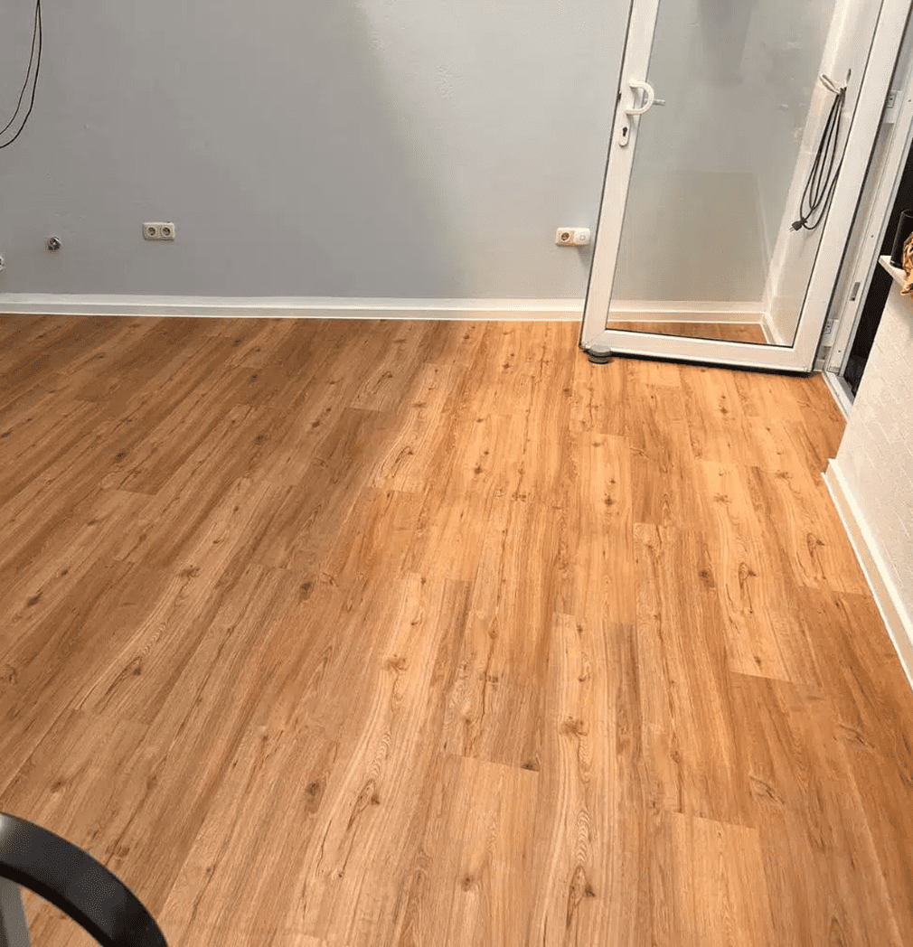 Ein Raum mit hellbraunem Holzfußboden, weißer Verkleidung, hellgrauen Wänden, mehreren Steckdosen an der Wand und einer Glastür, die teilweise nach draußen führt.