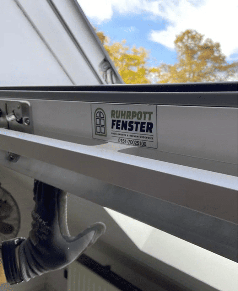 Eine behandschuhte Hand schiebt ein Fenster auf. Auf einem Schild am Fensterrahmen steht "RUHRPOTT FENSTER", darunter eine Telefonnummer. Draußen sind gelbe Herbstbäume zu sehen.