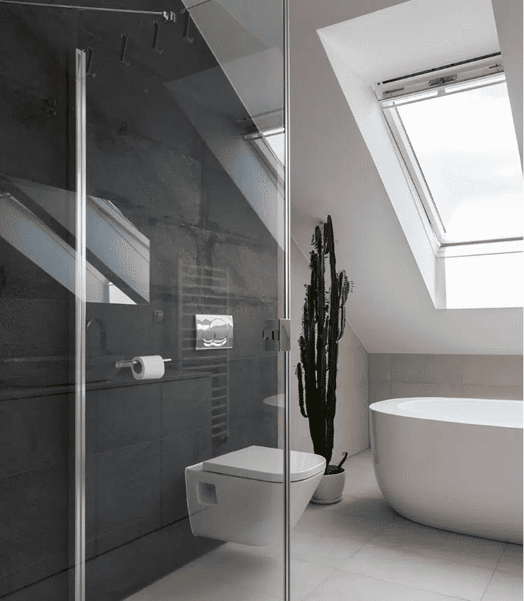 Modernes Badezimmer mit Glasdusche, wandmontierter Toilette, weißer Badewanne, Topfkakteen und großen, schrägen Dachfenstern, die natürliches Licht hereinlassen.