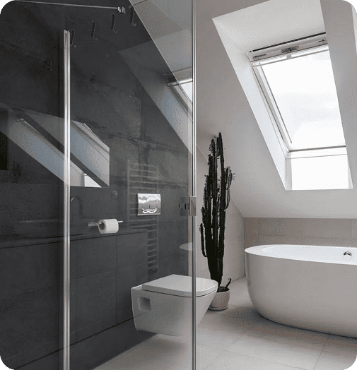 Modernes Badezimmer mit freistehender Badewanne, wandmontierter Toilette, dunkel gefliester Wand, Glasduschabtrennung, Topfkakteen und einem großen, schrägen Dachfenster, das natürliches Licht hereinlässt.