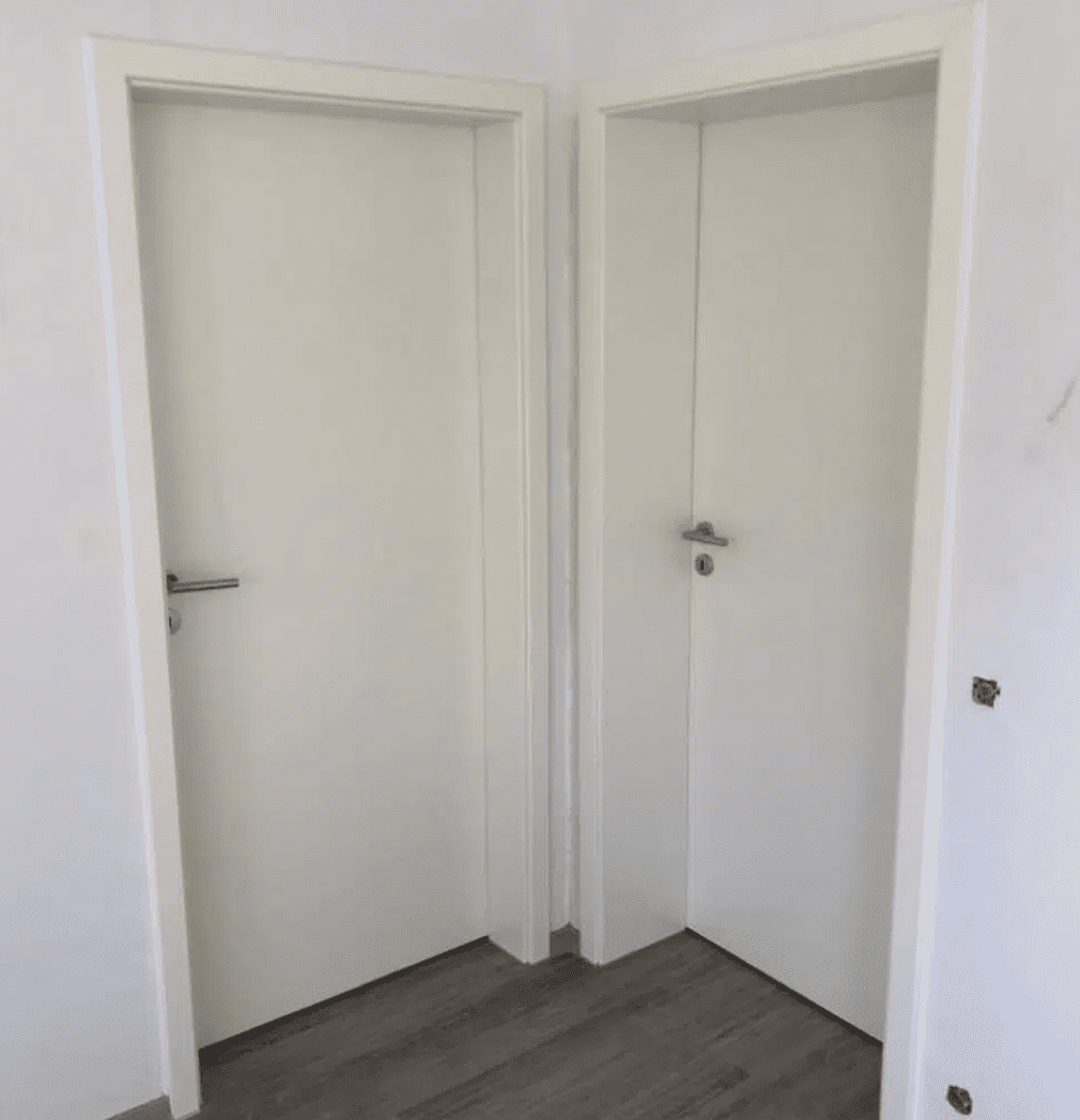 In einem Raum mit weißen Wänden und holzgemusterten Fußböden stehen zwei weiße Türen mit silbernen Griffen dicht nebeneinander in einer Ecke.