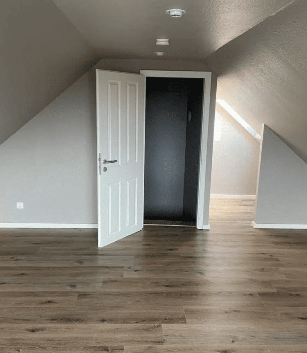 Ein leerer Dachraum mit hellgrauen Wänden, einer schrägen Decke, Holzfußboden, einer offenen weißen Tür und einem kleinen Fenster, das Tageslicht hereinlässt.