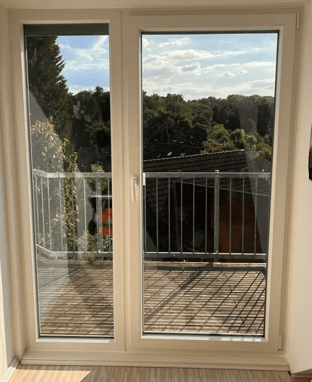 Eine Glastür öffnet sich zu einem kleinen Balkon mit einem Metallgeländer. Draußen sind Bäume, Dächer und ein teilweise bewölkter Himmel zu sehen, wobei das Sonnenlicht Schatten auf den Balkonboden wirft.