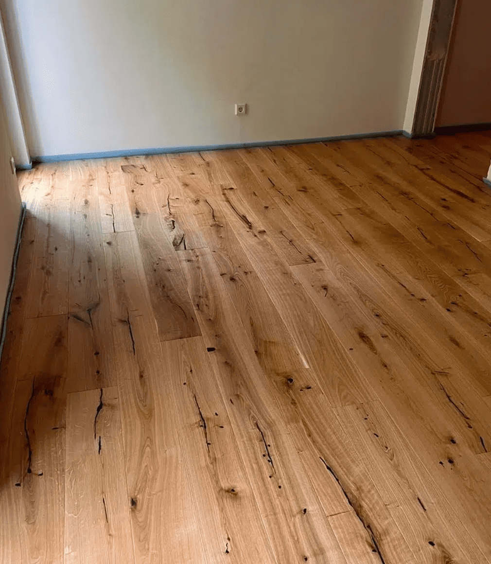 Ein Raum mit frisch verlegtem hellbraunem Holzfußboden, der sichtbare natürliche Maserungen und Äste aufweist, mit cremefarbenen Wänden und einer Steckdose an der gegenüberliegenden Wand. Der Raum ist leer und gut beleuchtet.