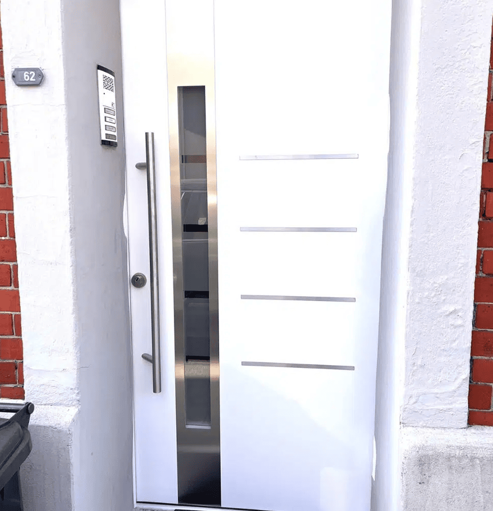 Eine moderne weiße Haustür mit einer vertikalen Glasscheibe und einem langen Metallgriff, eingefasst in einen weißen Rahmen mit einer Backsteinmauer auf der linken Seite. In der Nähe sind eine Türklingel und eine Sprechanlage angebracht, und die Hausnummer 62 ist sichtbar.