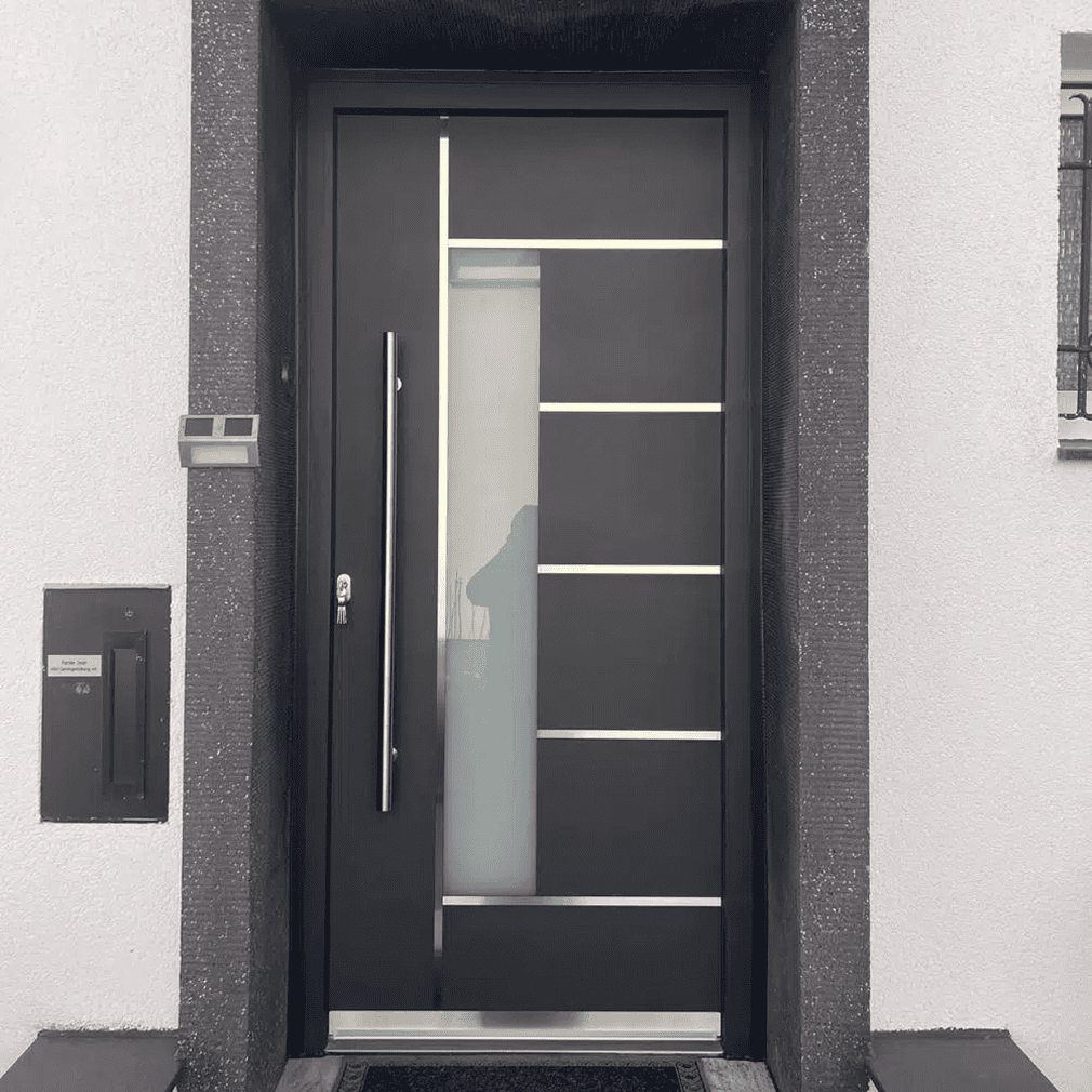 Moderne schwarze Haustür mit horizontalen silbernen Linien und einer vertikalen Milchglasscheibe, eingefasst in eine weiße Wand mit einem schmalen Briefschlitz und einer Türklingel auf der linken Seite.
