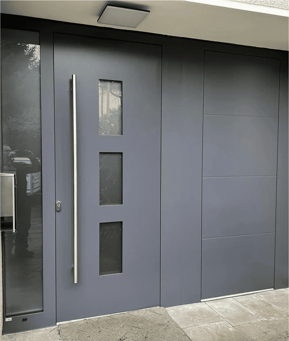 Eine moderne graue Haustür mit einem hohen silbernen Griff und drei rechteckigen Glasscheiben, daneben eine passende graue Wand und ein Pflaster.