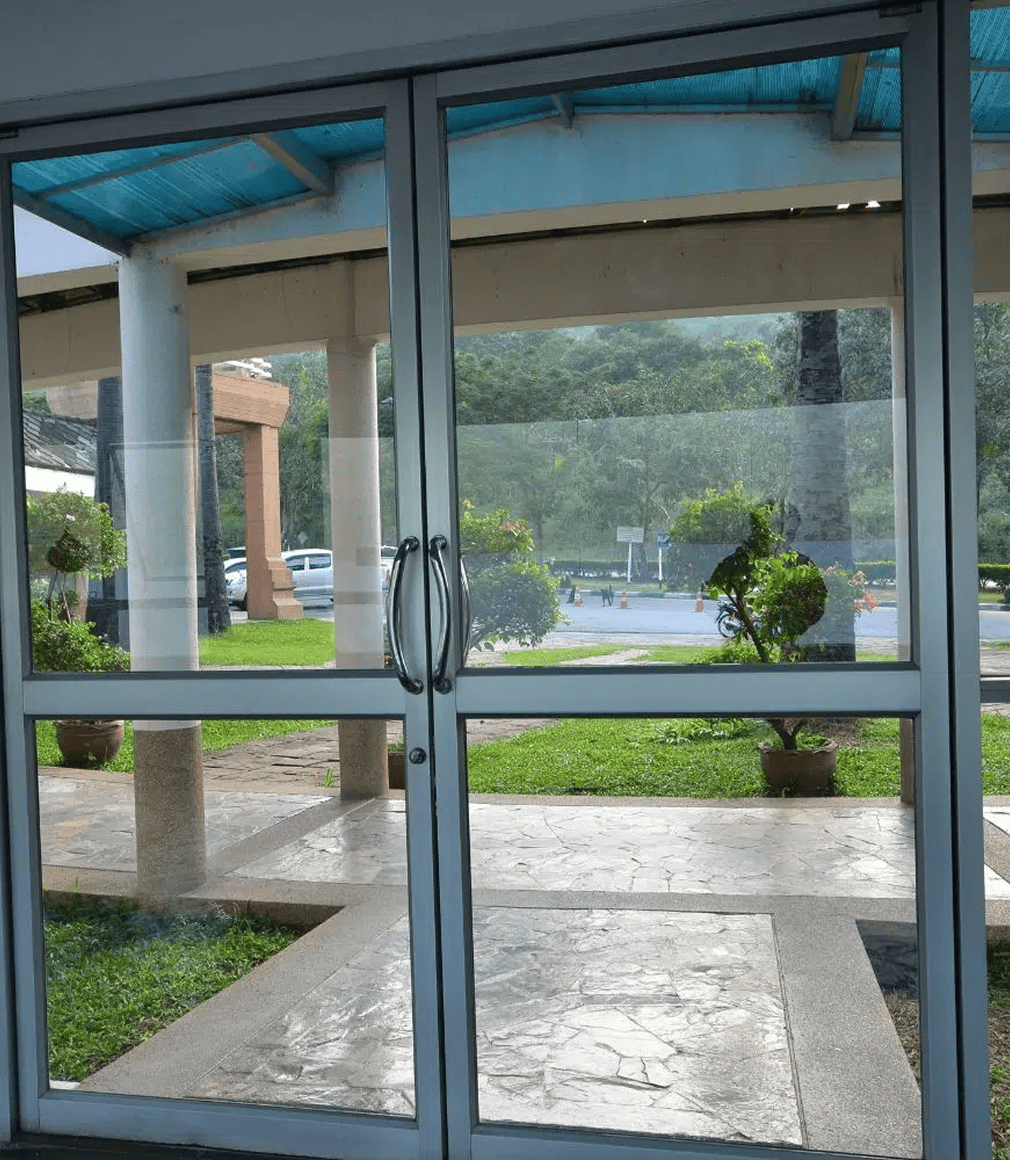 Ein Paar Doppeltüren aus Glas mit Metallgriffen öffnet sich zu einem überdachten Weg und einem Gartenbereich mit grünem Gras, großen Topfpflanzen und Bäumen. Der Himmel scheint bewölkt zu sein.