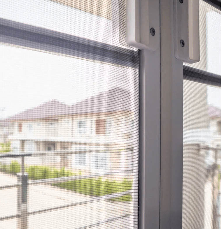 Nahaufnahme eines Fensters mit einem Maschendrahtgitter, das auf ein Wohnviertel mit Häusern und Grünflächen im Hintergrund hinausgeht. Das Bild ist aus dem Inneren eines Gebäudes aufgenommen.