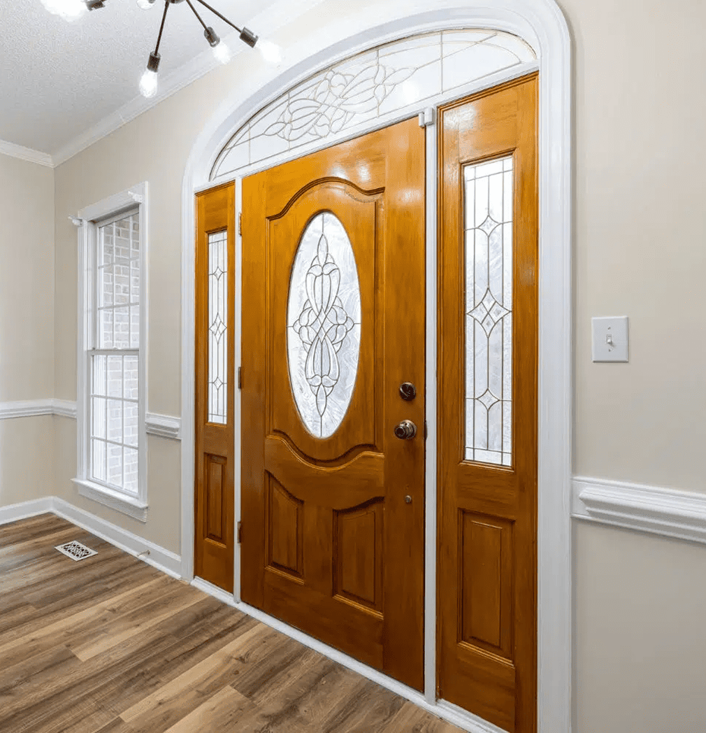 Eine hölzerne Eingangstür mit dekorativen Glasscheiben und einem gewölbten Oberlicht, umrahmt von einer weißen Zierleiste, in einem hellen Eingangsbereich mit beigen Wänden und Fußboden in Holzoptik.