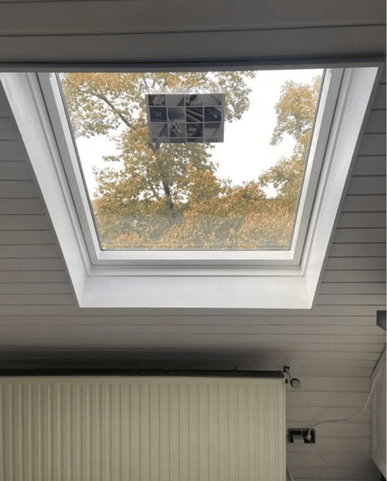 Ein Oberlichtfenster in einer weiß getäfelten Decke zeigt Baumzweige mit gelben Blättern im Freien. In der Mitte des Fensters ist ein kleines Objekt mit Fächern angebracht.