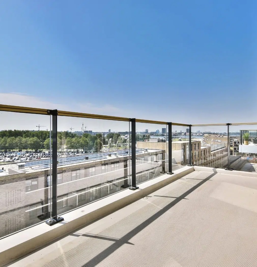 Sonnige Dachterrasse mit Glasgeländer und Blick auf eine Stadtlandschaft, einen Parkplatz und grüne Bäume unter einem klaren blauen Himmel.