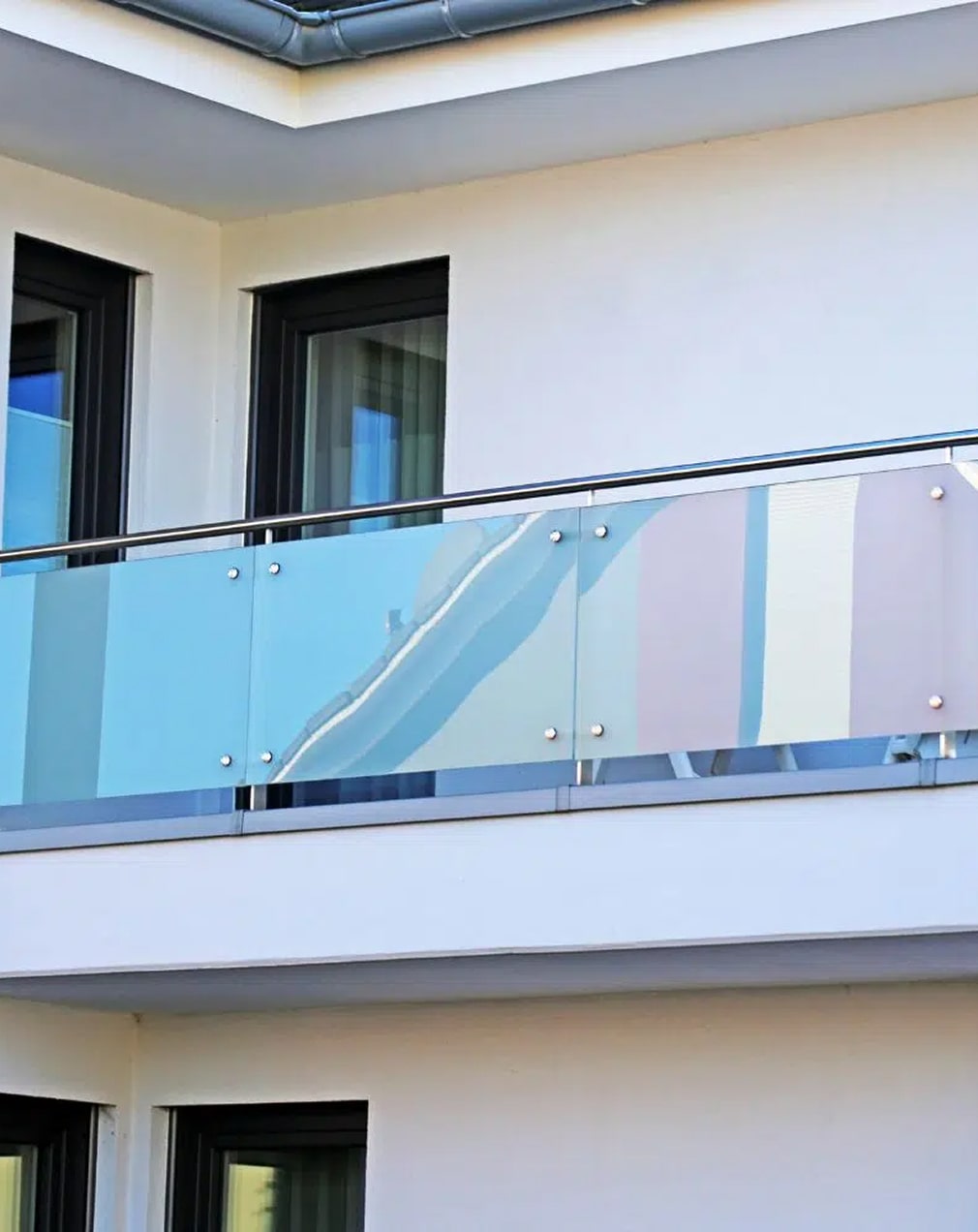 Ein modernes Wohnhaus mit einem gläsernen Balkongeländer, in dem sich der Himmel und die nahegelegenen Strukturen spiegeln und das vor großen Fenstern mit dunklen Rahmen steht.