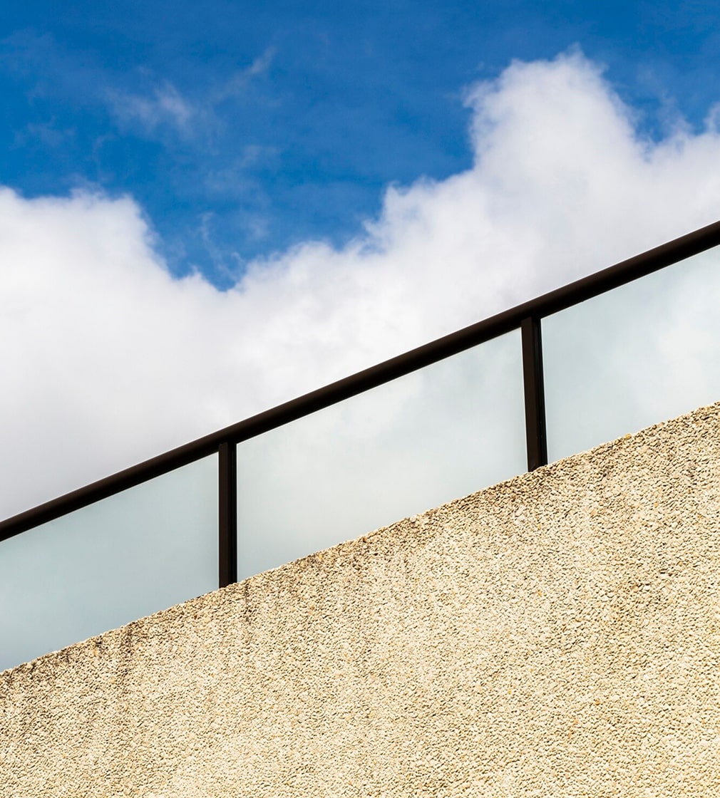 Eine beigefarbene, strukturierte Wand mit einem Geländer aus Glas und Metall vor einem blauen Himmel mit vereinzelten weißen Wolken.