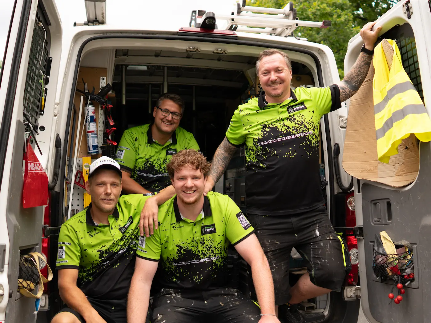Vier lächelnde Männer in grün-schwarzen Arbeitsuniformen posieren auf der Ladefläche eines mit Werkzeugen und Ausrüstung gefüllten Arbeitswagens. Zwei von ihnen sitzen und zwei stehen, wobei ein Arm auf der Wagentür ruht.
