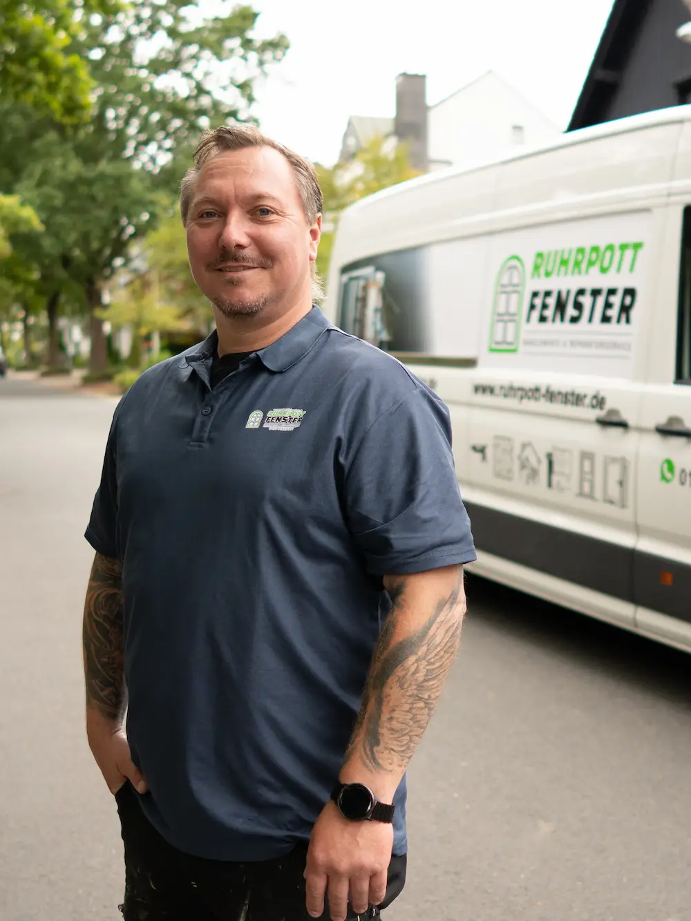 Ein Mann in einem marineblauen Poloshirt steht auf einer Straße und lächelt in die Kamera. Hinter ihm steht ein weißer Lieferwagen mit "Ruhrpott Fenster"-Schriftzug und Logo, der in der Nähe von Bäumen und Häusern geparkt ist.