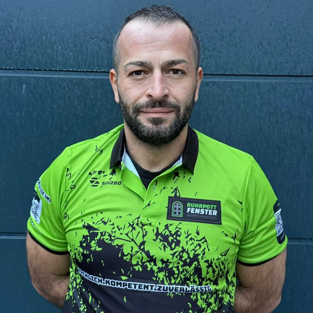 Ein Mann mit kurzen dunklen Haaren und einem Bart steht vor einer blauen Wand und trägt ein hellgrünes und schwarzes Sporttrikot mit verschiedenen Logos und Mustern.